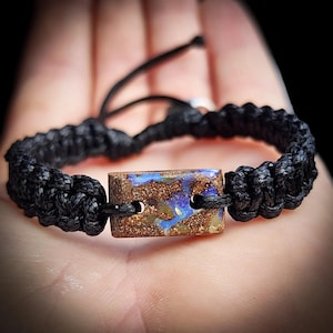 May include: Black macramé bracelet with a rectangular opal stone displaying blue and brown tones. The adjustable bracelet has a knotted design with a sliding knot closure. The opal stone is the central feature.