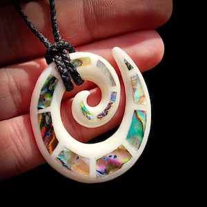 Op de afbeelding: Een spiraalvormige hanger van glad wit materiaal, voorzien van ingelegde iriserende abalone-schelpsegmenten in tinten groen, blauw en paars. De hanger hangt aan een zwart gevlochten koord en heeft een haakachtig ontwerp.