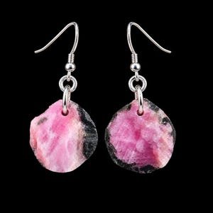 May include: A pair of drop earrings featuring raw, circular slices of pink and black tourmaline gemstone. Each stone is suspended from a silver-toned metal hook wire by a small circular jump ring, showcasing a natural, textured surface.