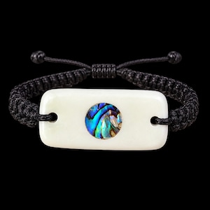 May include: A bracelet featuring a rectangular off-white bone-colored center piece with a circular iridescent abalone shell inlay at the middle. The center piece is attached to a black woven cord band with adjustable sliding knots at the ends.