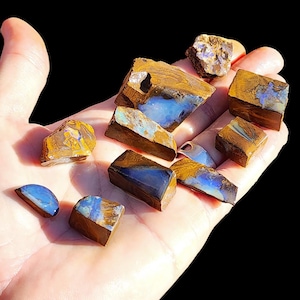Puede incluir: Una colección de diez especímenes de ópalo boulder en bruto con matriz de piedra de hierro marrón y destellos iridiscentes azules y verdes. Las piedras varían en forma y tamaño, mostrando texturas naturales y superficies pulidas que reflejan la luz.