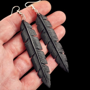 Könnte beinhalten: Ein Paar lange, schwarze, federförmige Ohrringe mit detaillierten, schnitzartigen Texturen auf der Oberfläche. Jeder Ohrring verfügt über einen silberfarbenen Metallhaken zur Befestigung, wodurch ein hängendes Design entsteht.