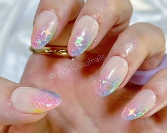 Puede incluir: Primer plano de una mano con uñas ovaladas pintadas con un degradado de colores pastel, incluyendo rosa, azul y amarillo. Cada uña está adornada con una pequeña estrella iridiscente.