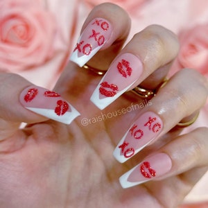 Puede incluir: U&ntilde;as artificiales largas en forma de ata&uacute;d con un dise&ntilde;o de punta francesa blanca. Las u&ntilde;as presentan acentos de purpurina roja de labios y el texto "xo xo". Las u&ntilde;as est&aacute;n sobre un fondo rosa suave.
