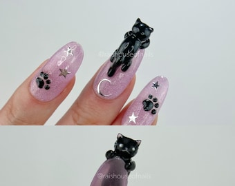 Uñas postizas 3D con diseño de gato negro, huellas de patas y estrellas plateadas.