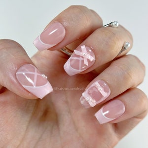 Handmade pink coquette press on nails with sheer pink ribbon bows, white cross-hatch ribbon pattern, pink French tips, and pearl accents on a sheer pink gel base, shown in coffin extra short by Rai's House of Nails
