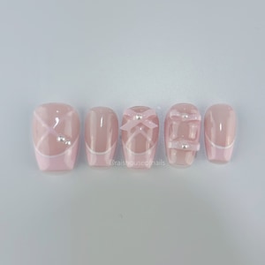 May include: A set of five pink press-on nails. Each nail features a different design, including white accents, bows, and small silver embellishments. The nails have a glossy finish and a square shape. The text "@raishouseofnails" is visible.