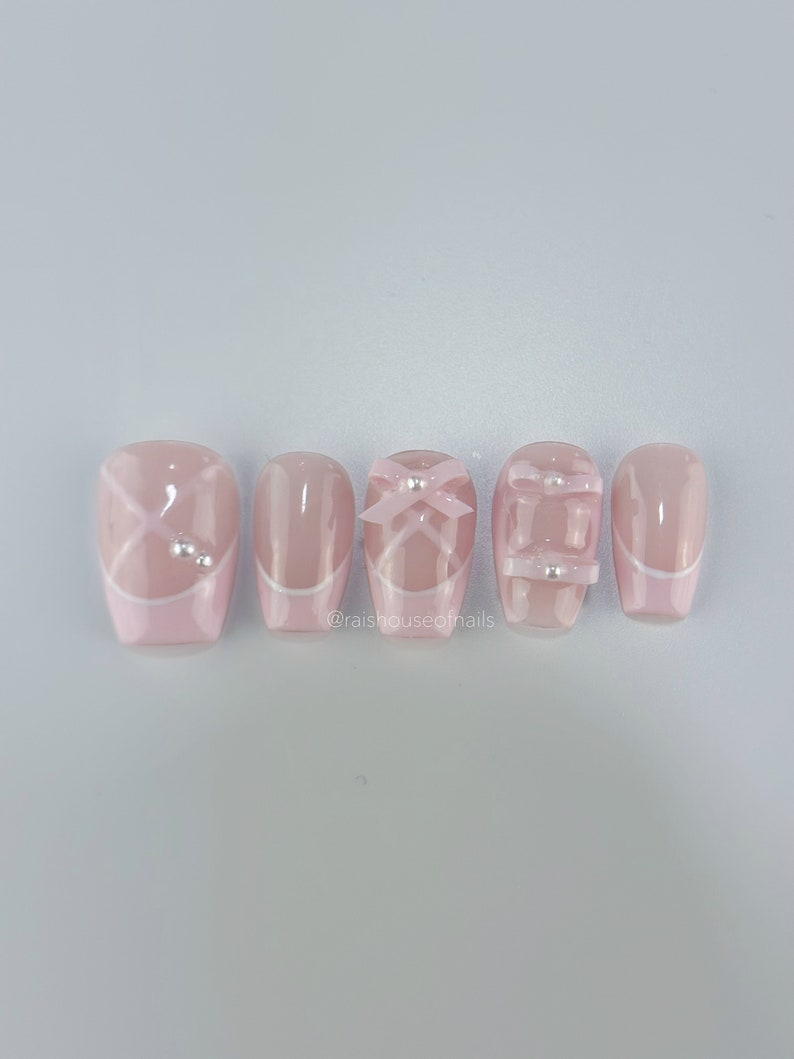 May include: A set of five pink press-on nails. Each nail features a different design, including white accents, bows, and small silver embellishments. The nails have a glossy finish and a square shape. The text "@raishouseofnails" is visible.