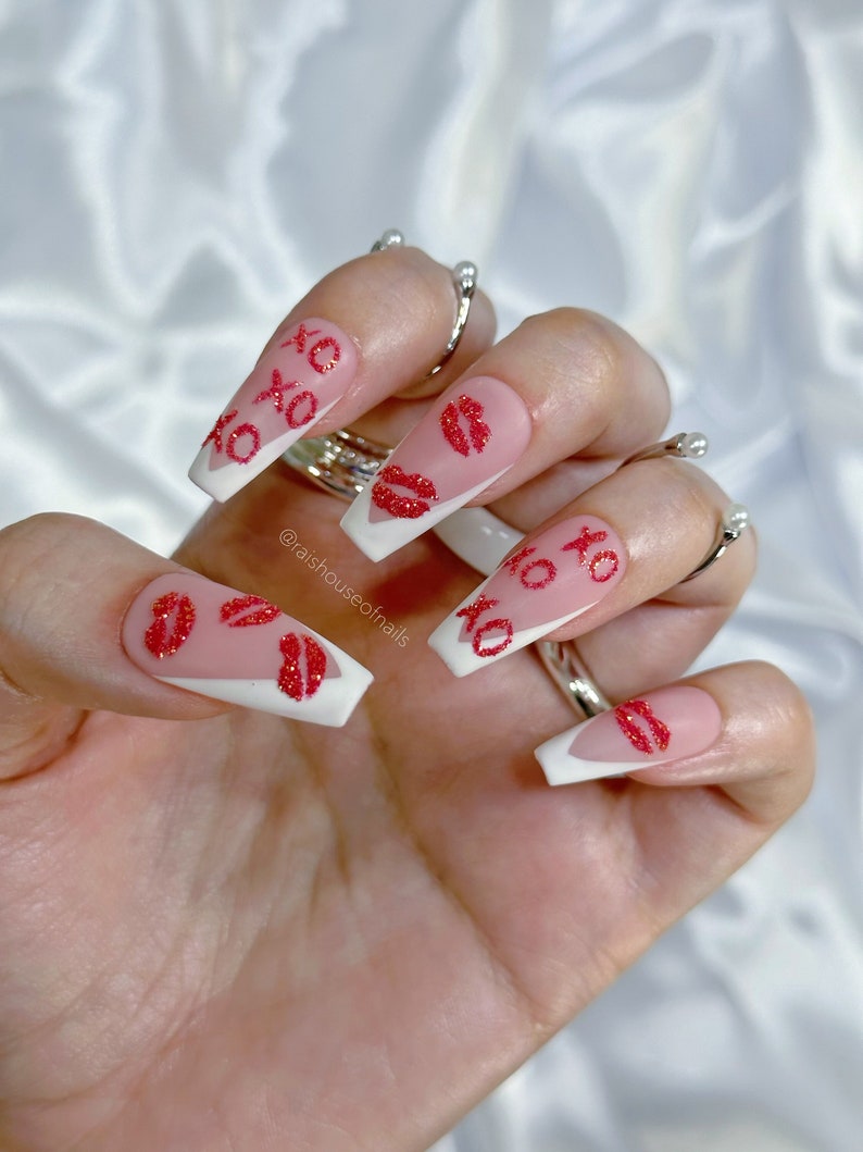 Handmade XOXO glitter press on nails with red glitter XOXO lettering, red glitter kiss marks, and white V-shaped French tips on a pink nude gel base, shown in coffin medium by Rai's House of Nails