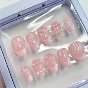 May include: A set of ten oval-shaped, light pink press-on nails. Each nail features a white French tip design and some nails have a bow or gem embellishments. The nails are displayed in a light blue case.