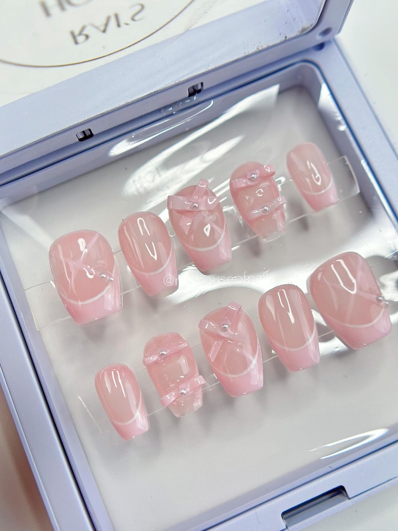 May include: A set of ten oval-shaped, light pink press-on nails. Each nail features a white French tip design and some nails have a bow or gem embellishments. The nails are displayed in a light blue case.