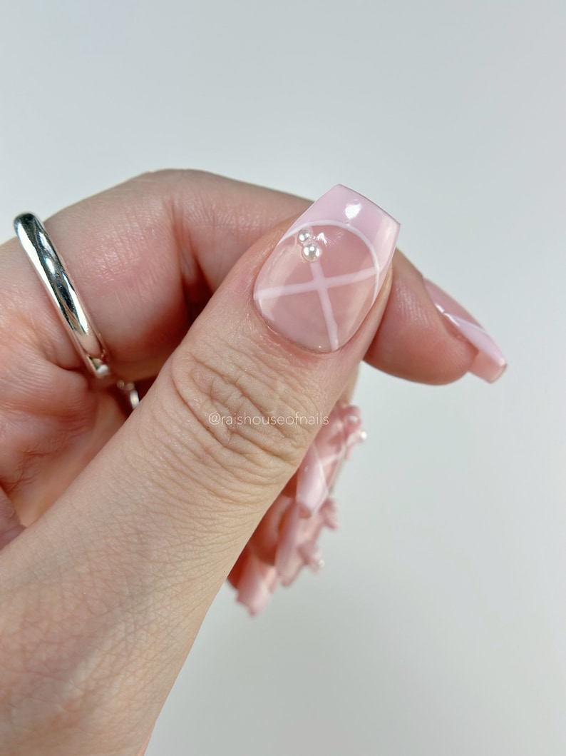 May include: Close-up of a hand with a silver ring and long, square-shaped artificial nails. The nails are painted light pink with white geometric designs and small rhinestones. The image is set against a white background.