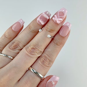 May include: A set of artificial nails with a French manicure design. The nails are a light pink with white tips and feature white ribbon and pearl accents. The nails are shown on a hand with silver rings.