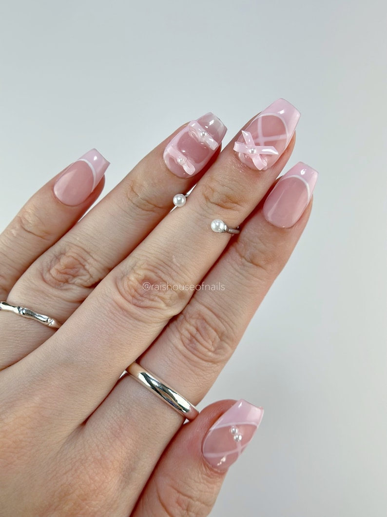 May include: A set of artificial nails with a French manicure design. The nails are a light pink with white tips and feature white ribbon and pearl accents. The nails are shown on a hand with silver rings.