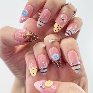 Unhas postiças kawaii com ursinhos de pelúcia, pingentes de botões e pontas listradas.