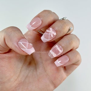 May include: A hand displaying a set of pink and white artificial nails. The nails feature a French tip design, white crisscross patterns, and small bow accents. The nails are square-shaped and have a glossy finish. A silver ring with pearl accents is also visible.