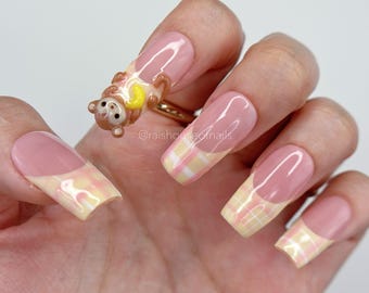 Puede incluir: Una mano con uñas artificiales largas y cuadradas. Las uñas presentan un diseño de cuadros rosas y amarillos en las puntas y una base rosa. También se ve un pequeño anillo de oso marrón con una luna creciente amarilla.