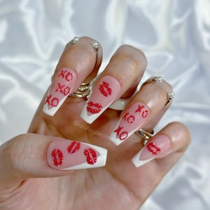 Puede incluir: U&ntilde;as acr&iacute;licas largas en forma de ata&uacute;d con base rosa, puntas blancas y dise&ntilde;os de besos y XO con purpurina roja. Las u&ntilde;as est&aacute;n adornadas con anillos plateados. La imagen est&aacute; sobre un fondo blanco y sedoso.