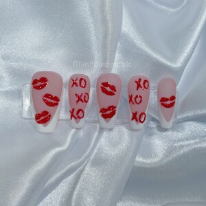 Puede incluir: Un conjunto de cinco u&ntilde;as artificiales con base rosa claro y puntas blancas. Cada u&ntilde;a presenta marcas de besos de purpurina roja y las letras "XO" en rojo. Las u&ntilde;as est&aacute;n dispuestas sobre un soporte de pl&aacute;stico transparente.
