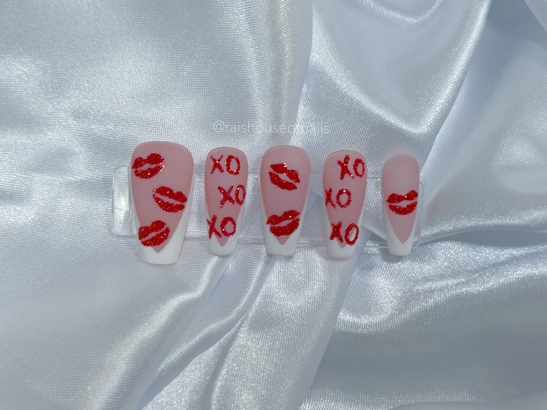 Puede incluir: Un conjunto de cinco u&ntilde;as artificiales con base rosa claro y puntas blancas. Cada u&ntilde;a presenta marcas de besos de purpurina roja y las letras "XO" en rojo. Las u&ntilde;as est&aacute;n dispuestas sobre un soporte de pl&aacute;stico transparente.