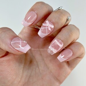 May include: A hand with a manicure featuring pink and white French tips. Some nails have white crisscross designs, bows, and pearl accents. The nails are square-shaped and have a glossy finish. The manicure is elegant and feminine.