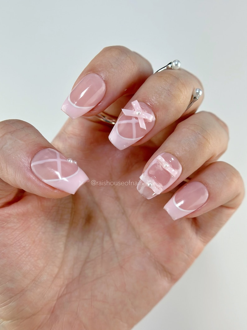 May include: A hand with a manicure featuring pink and white French tips. Some nails have white crisscross designs, bows, and pearl accents. The nails are square-shaped and have a glossy finish. The manicure is elegant and feminine.