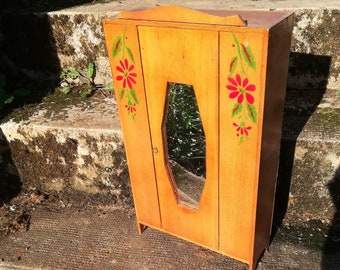 May include: A small wooden cabinet with a mirror door and painted floral details. The cabinet is painted in a light wood tone with red and green flowers.