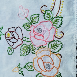 Könnte beinhalten: Weiße Leinen-Tischdecke mit gesticktem Blumendesign. Das Design zeigt rosa, orange und braune Rosen mit grünen Blättern.
