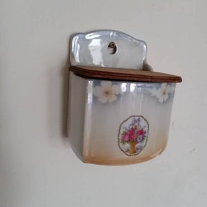 May include: A white ceramic wall mounted salt or sugar dispenser with a floral design and a wooden lid.