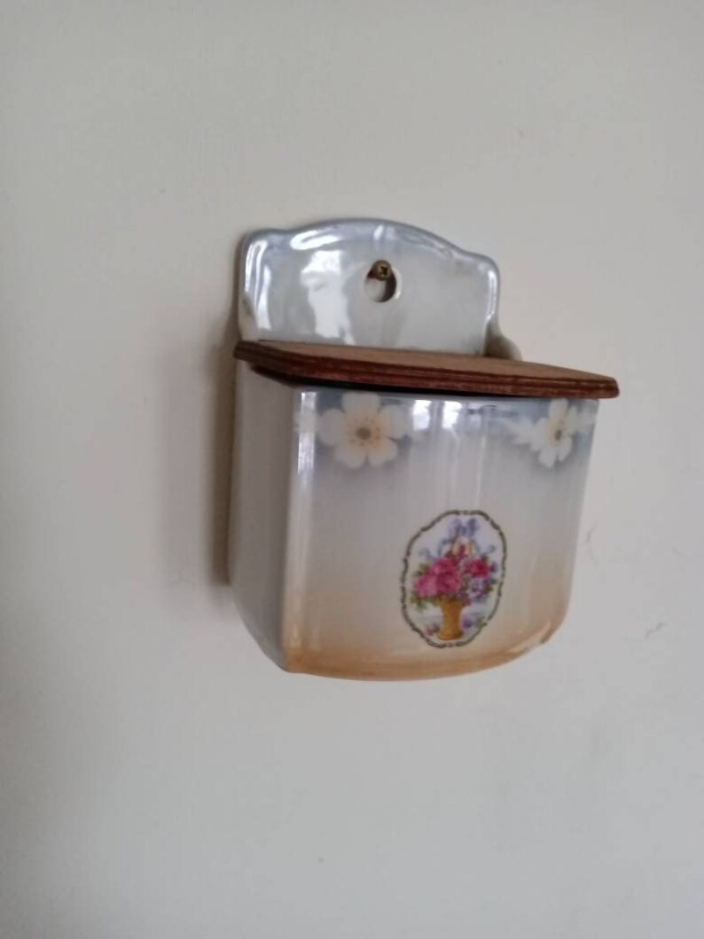May include: A white ceramic wall mounted salt or sugar dispenser with a floral design and a wooden lid.