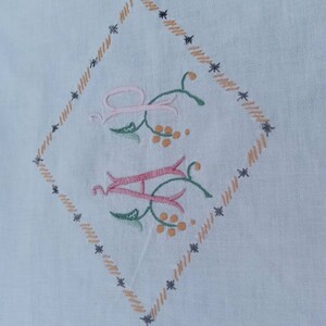 May include: A white fabric with a diamond-shaped embroidered design. The design features the letters "A" and "H" in pink and green with floral accents. The diamond is outlined with a brown and black stitching.