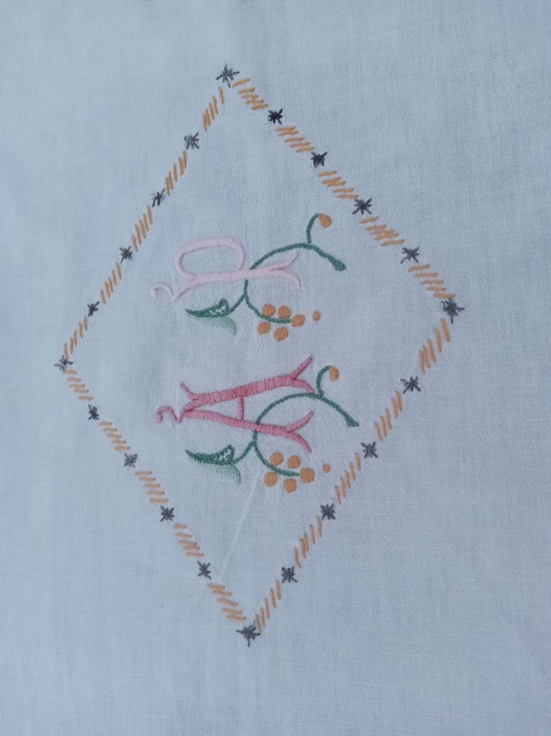 May include: A white fabric with a diamond-shaped embroidered design. The design features the letters "A" and "H" in pink and green with floral accents. The diamond is outlined with a brown and black stitching.