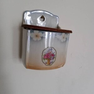 May include: A white ceramic wall mounted salt or sugar dispenser with a floral design and a wooden lid.