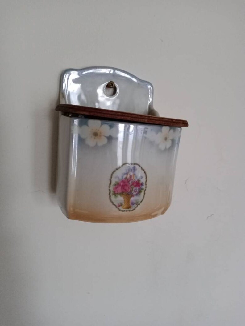 May include: A white ceramic wall mounted salt or sugar dispenser with a floral design and a wooden lid.