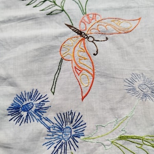 Peut inclure: Tissu blanc brodé avec un papillon coloré et des fleurs bleues. Le papillon a des ailes orange et jaunes avec des accents bruns. Les fleurs ont des pétales bleus et des tiges vertes.