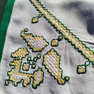 May include: Close-up of a white linen textile with embroidered floral patterns. The embroidery features yellow and white cross-stitch designs outlined in green. The edge of the textile is trimmed with a green border.