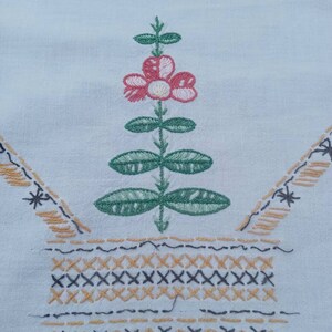May include: A white fabric with a hand-stitched design of a green plant with a pink flower in a brown and black basket.