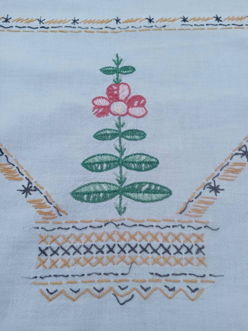 May include: A white fabric with a hand-stitched design of a green plant with a pink flower in a brown and black basket.