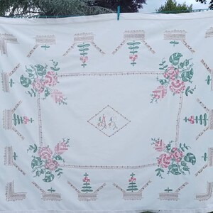 May include: White linen tablecloth with embroidered floral designs in pink and green. The centre features a diamond-shaped monogram with the initials "A.A."