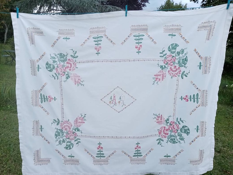 May include: White linen tablecloth with embroidered floral designs in pink and green. The centre features a diamond-shaped monogram with the initials "A.A."