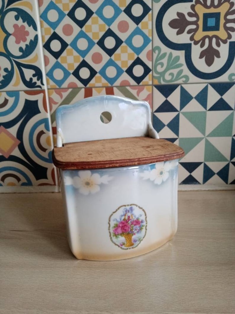 May include: A vintage white ceramic wall mounted sugar dispenser with a wooden lid and a floral design. The dispenser has a blue and white glaze and a floral pattern with pink flowers and green leaves.