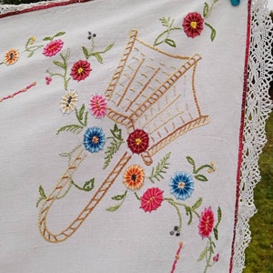 Puede incluir: Mantel de mesa de lino blanco con flores bordadas y un diseño de cesta. La cesta está bordada con hilo marrón y las flores son de color rosa, rojo, naranja, amarillo, azul y verde.