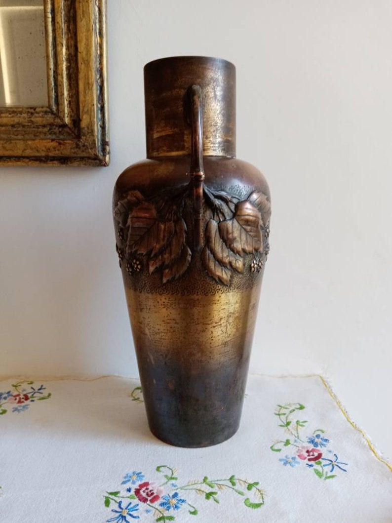 Op de afbeelding: Een hoge, antieke bronzen vaas met een getextureerd oppervlak en reli&euml;fmotieven van bladeren. De vaas heeft twee handvatten en een licht uitlopende opening.