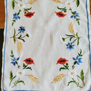 May include: White linen tablecloth with embroidered flowers in red, blue, white and yellow. The flowers are arranged in a border around the edge of the tablecloth.