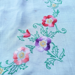 Puede incluir: Un mantel azul claro con enredaderas verdes bordadas y flores rosas, moradas y blancas.