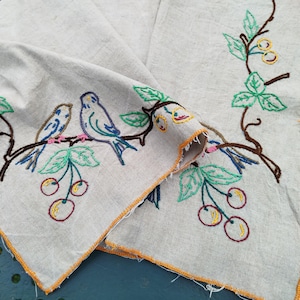 May include: A white linen tablecloth with embroidered birds and cherries. The birds are blue, green and yellow, and the cherries are red and yellow. The tablecloth has a yellow border.