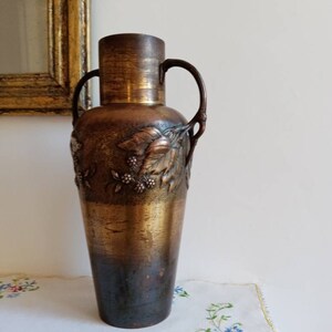 Op de afbeelding: Een hoge, antieke bronzen vaas met een getextureerd oppervlak en een bloemenmotief. De vaas heeft twee handvatten en een licht uitlopende bovenkant.