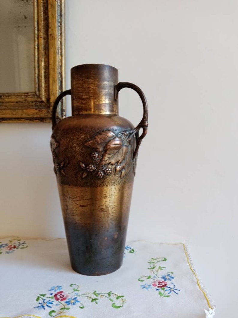Op de afbeelding: Een hoge, antieke bronzen vaas met een getextureerd oppervlak en een bloemenmotief. De vaas heeft twee handvatten en een licht uitlopende bovenkant.
