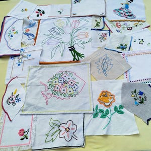 Op de afbeelding: Een verzameling van diverse witte stoffen vierkanten en rechthoeken met verschillende kleurrijke bloemen- en decoratieve borduurontwerpen, waaronder tulpen, rozen en een figuur in een jurk met ruches, met sommige stukken met schulpranden of franjes.