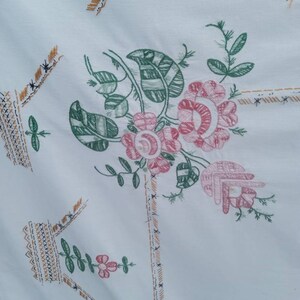 May include: White tablecloth with an embroidered floral design. The flowers are pink and green with a woven pattern. The tablecloth has a border of brown and gold stitching.